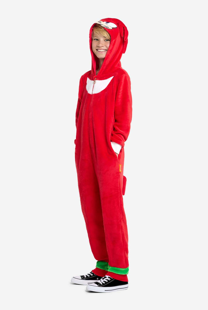 A kid is wearing a bright red Knuckles the Echidna-themed kids onesie from OppoSuits. The fun and cozy onesie features a zip-up front and an attached hood designed to resemble Knuckles' face, complete with expressive eyes and floppy dreadlocks. The suit is primarily red, with a white crescent pattern on the chest, reflecting Knuckles' iconic look from the Sonic the Hedgehog video game series. 