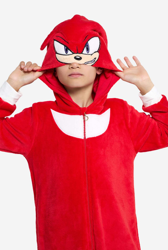 A kid is wearing a bright red Knuckles the Echidna-themed kids onesie from OppoSuits. The fun and cozy onesie features a zip-up front and an attached hood designed to resemble Knuckles' face, complete with expressive eyes and floppy dreadlocks. The suit is primarily red, with a white crescent pattern on the chest, reflecting Knuckles' iconic look from the Sonic the Hedgehog video game series. 