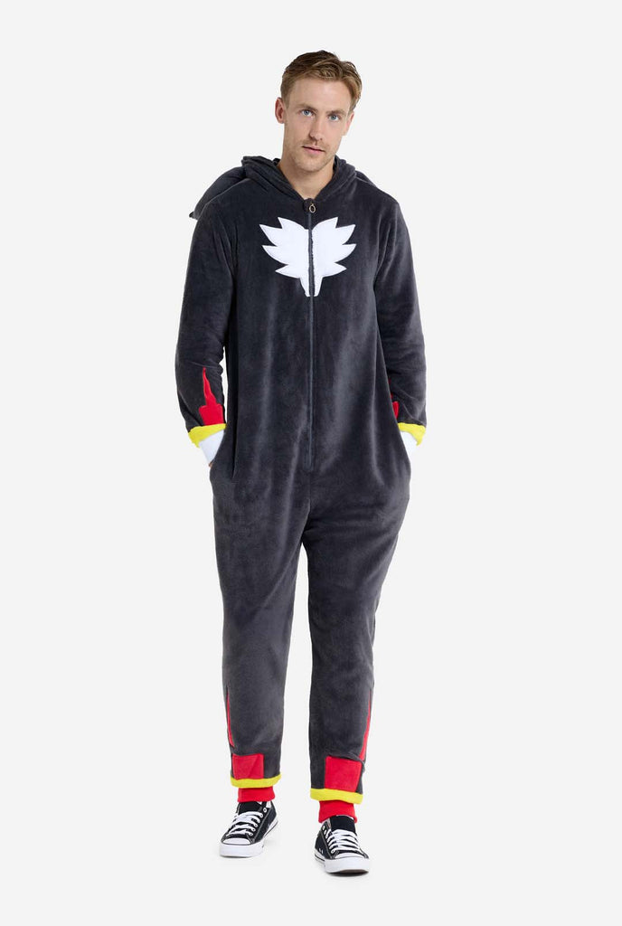 A man wearing a plush black onesie inspired by the popular hedgehog character Shadow, featuring spiky red and black accents on the hood, a white chest emblem, and yellow wrist details. The onesie is made from soft fleece material, offering comfort and a playful design for cosplay, lounging, or themed events. The person smiles while the hood is lying on his back. Perfect for fans of gaming or animated characters. Full body image.