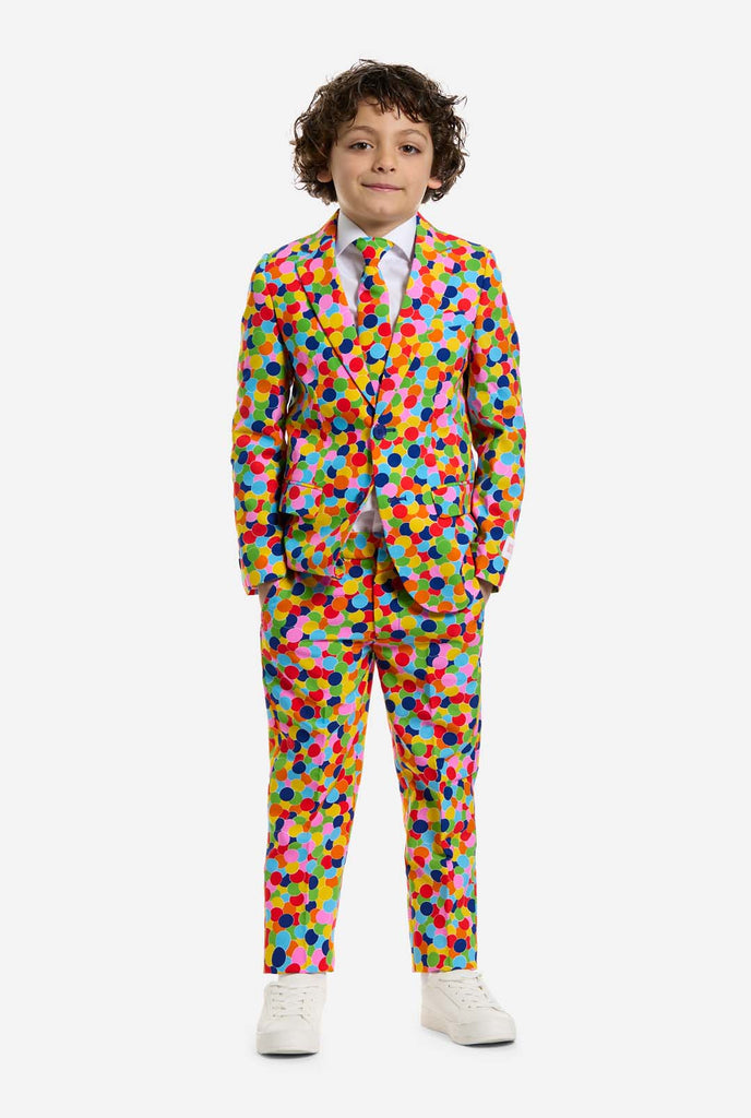 In this image, a kid is wearing a boys’ suit with all-over multicolor confetti print and white shirt. The suit has a bright pattern with circles in various colors including orange, yellow, pink, green, blue, and red.