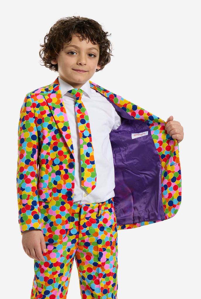 In this image, a kid is wearing a boys’ suit with all-over multicolor confetti print and white shirt. The suit has a bright pattern with circles in various colors including orange, yellow, pink, green, blue, and red. Holding one side of the jacket open.