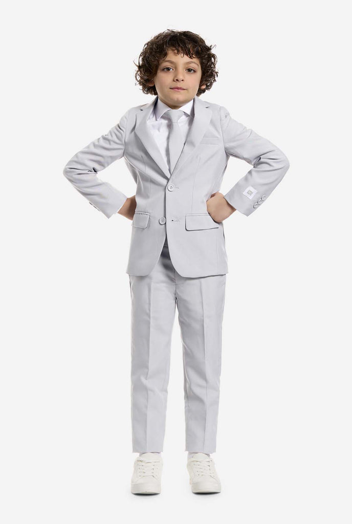 In this image, a kid is wearing a grey formal boys’ suit with a matching tie and a white shirt. The suit consists of a jacket, pants and clip on tie.