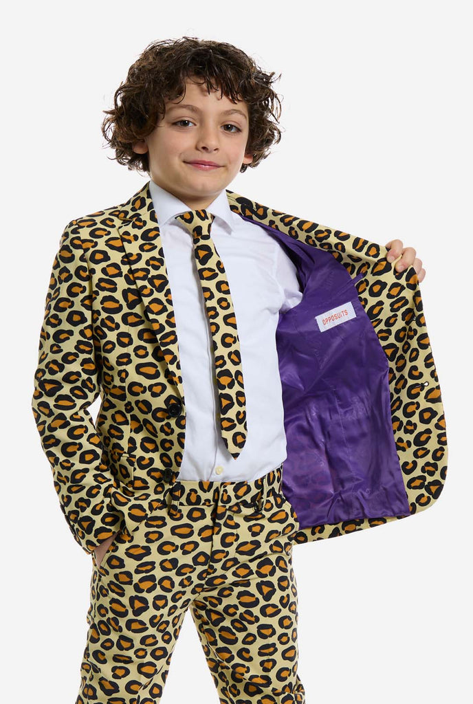 In this image, a kid is wearing a boys’ suit with all-over panther print and white shirt. The suit is beige with brown and black spotted panther print. Suits consists of jacket, pants and clip-on tie. Holding one side of the jacket open.