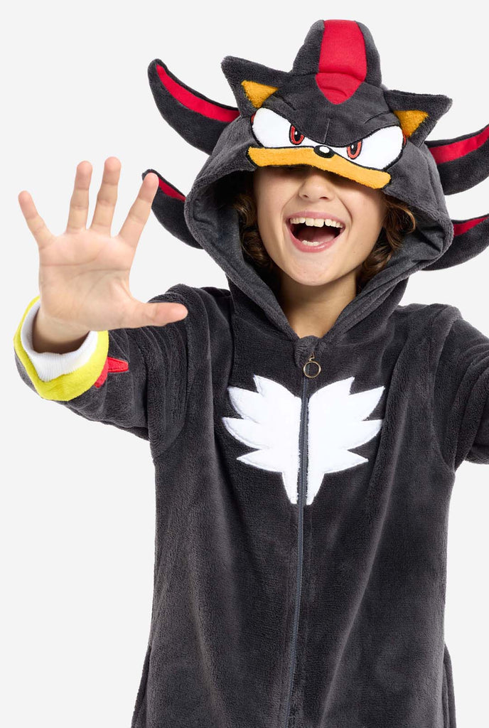 A kid wearing a plush black kids onesie inspired by the popular hedgehog character Shadow, featuring spiky red and black accents on the hood, a white chest emblem, and yellow wrist details. The onesie is made from soft fleece material, offering comfort and a playful design for cosplay, lounging, or themed events. Close up with the hood over the eyes. Perfect for fans of gaming or animated characters.