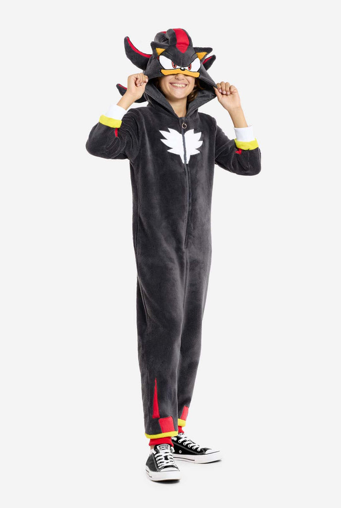 A kid wearing a plush black kids onesie inspired by the popular hedgehog character Shadow, featuring spiky red and black accents on the hood, a white chest emblem, and yellow wrist details. The onesie is made from soft fleece material, offering comfort and a playful design for cosplay, lounging, or themed events. The person smiles while adjusting the hood, showcasing the character's recognizable face and bold design. Perfect for fans of gaming or animated characters.