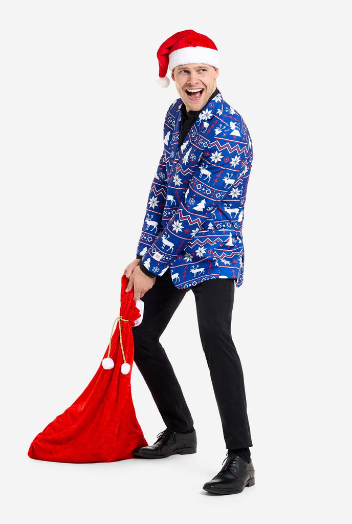 Man wearing blue Christmas blazer for men with Nordic Christmas figures. Man also wearing a black pants and black men's shirt, 
Christmas hat and carrying Christmas bag.