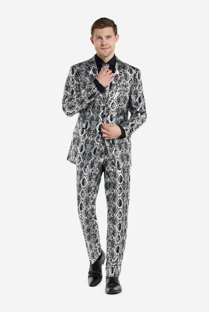 A man is wearing an eye-catching silver and black snake-print suit by OppoSuits and black shoes. The striking suit features a bold snakeskin pattern, offering a sleek, fashionable look perfect for parties, events, or making a daring fashion statement. The men suit is paired with a black dress shirt and a matching snake-print tie, adding a sophisticated edge to the overall ensemble. The man's confident smile and formal yet modern outfit give off a high-fashion, stylish vibe.