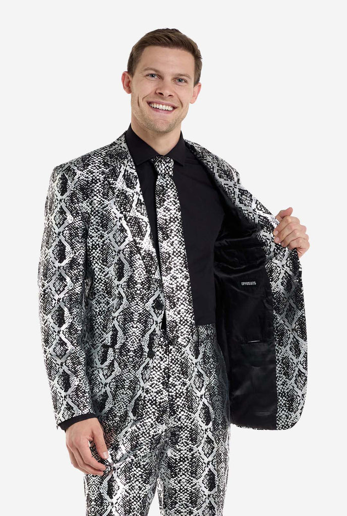 A man is wearing an eye-catching silver and black snake-print suit by OppoSuits. The striking suit features a bold snakeskin pattern, offering a sleek, fashionable look perfect for parties, events, or making a daring fashion statement. The men suit is paired with a black dress shirt and a matching snake-print tie, adding a sophisticated edge to the overall ensemble. The man is holding on side of the jacket open.