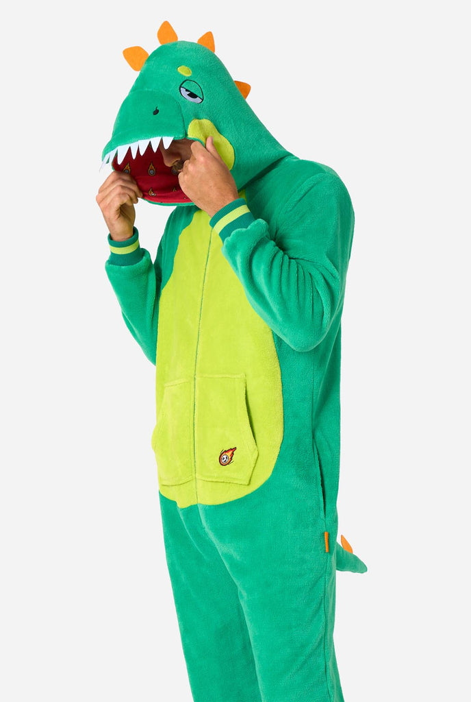 A man is wearing a green dinosaur adult onesie featuring playful hood details and cozy, soft material, perfect for lounging or costume events. He is standing sideways and holding the hood over his head.