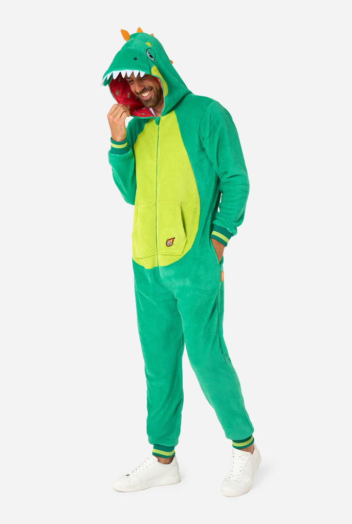 A man is wearing a green dinosaur adult onesie featuring playful hood details and cozy, soft material, perfect for lounging or costume events. He is standing sideways and holding the hood over his head with one hand. Full body image.