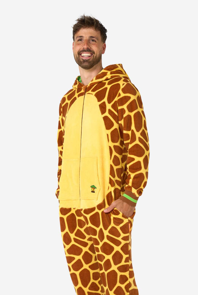 A smiling man is wearing a brown and yellow adult onesie designed as a giraffe. The onesie features a hood resembling a Giraffes head, and giraffe details, perfect for cozy lounging or costume parties. The man standing with his hands in the pockets.