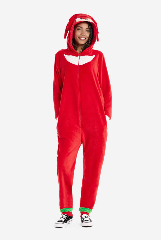 A woman wears a bright red Knuckles the Echidna adult onesie, a cozy and playful outfit inspired by the famous character from the Sonic the Hedgehog series. The onesie features Knuckles' signature white chest patch and includes a hood that showcases Knuckles' face, complete with his expressive eyes and long red dreads. The front zipper closure allows for easy wear, and the woman stands with hands I the side pockets.  The outfit, made from soft fleece material, is perfect for lounging, or cosplay.