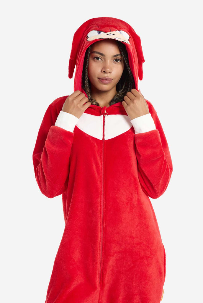 A woman wears a bright red Knuckles the Echidna adult onesie, a cozy and playful outfit inspired by the famous character from the Sonic the Hedgehog series. The onesie features Knuckles' signature white chest patch and includes a hood that showcases Knuckles' face, complete with his expressive eyes and long red dreads. The front zipper closure allows for easy wear, and the woman holding the hood. The outfit, made from soft fleece material, is perfect for lounging, cosplay, or themed events.