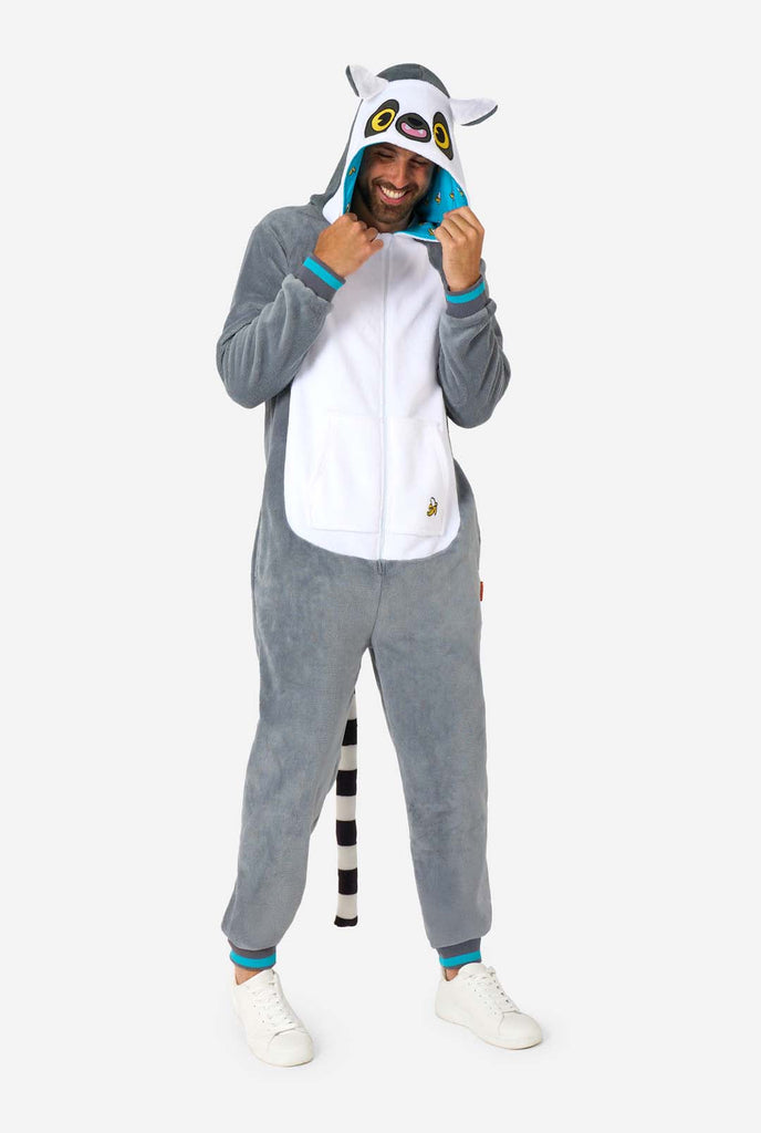 A man is wearing a grey and white lemur adult onesie featuring playful hood details and cozy, soft material, perfect for lounging or costume events. Including black and white tail. Full body image and the man is holding the hood.