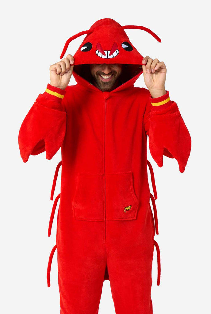 A man is wearing a bright red lobster-themed adult onesie featuring playful hood details and cozy, soft material, perfect for lounging or costume events. including stitched gloves as claws. Holding the hood over his eyes.