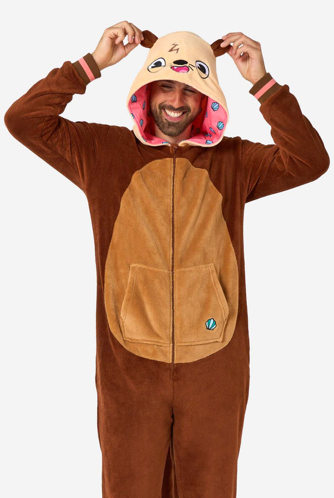 A smiling man is wearing a brown adult onesie designed as an otter. The onesie features a hood with ears, a cute face, and playful otter details, perfect for cozy lounging or costume parties. The man is holding the ears on the hood.