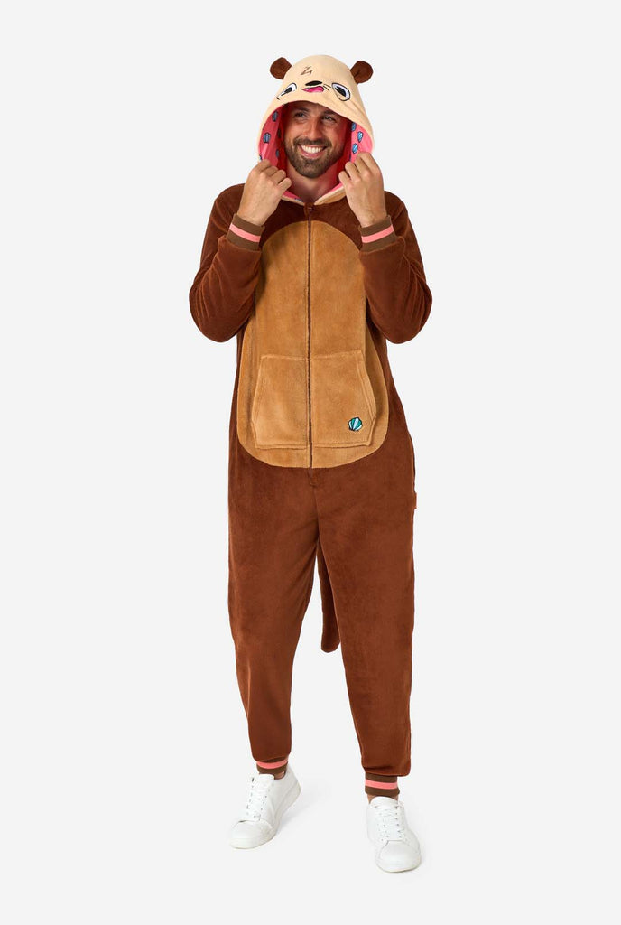 A smiling man is wearing a brown adult onesie designed as an otter. The onesie features a hood with ears, a cute face, and playful otter details, perfect for cozy lounging or costume parties. Full body image and the man is holding the hood.