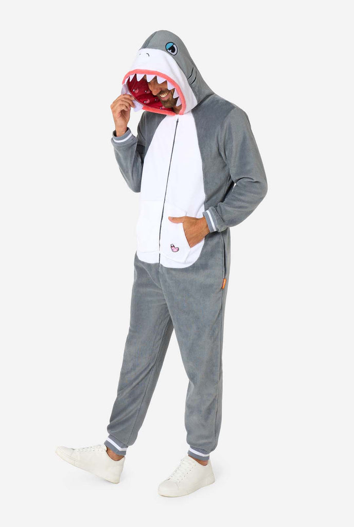 A man is wearing a grey and white shark adult onesie featuring playful hood details and cozy, soft material, perfect for lounging or costume events. Full body image and the man is holding the hood with one hand.