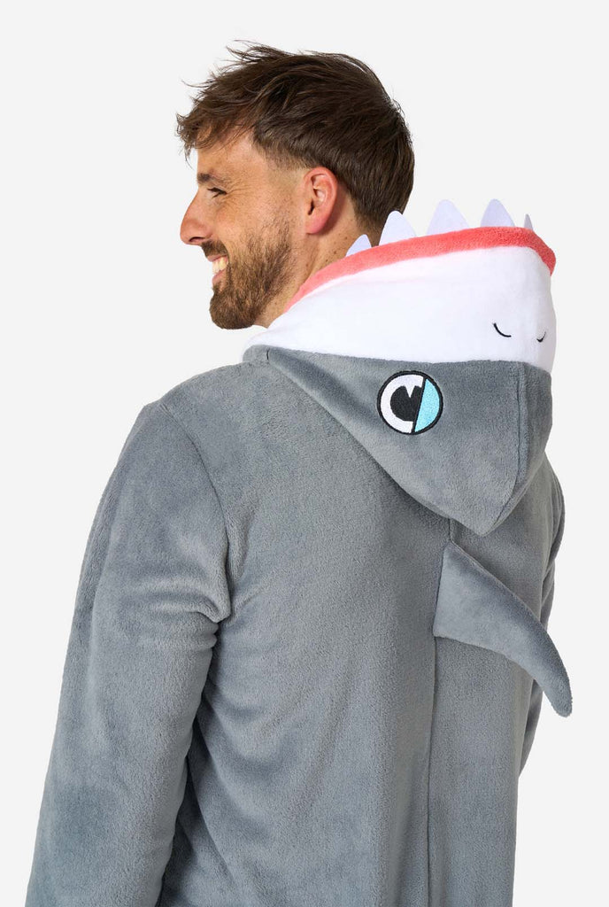 A man is wearing a grey and white shark adult onesie featuring playful hood details and cozy, soft material, perfect for lounging or costume events. Upper body close-up from the back.