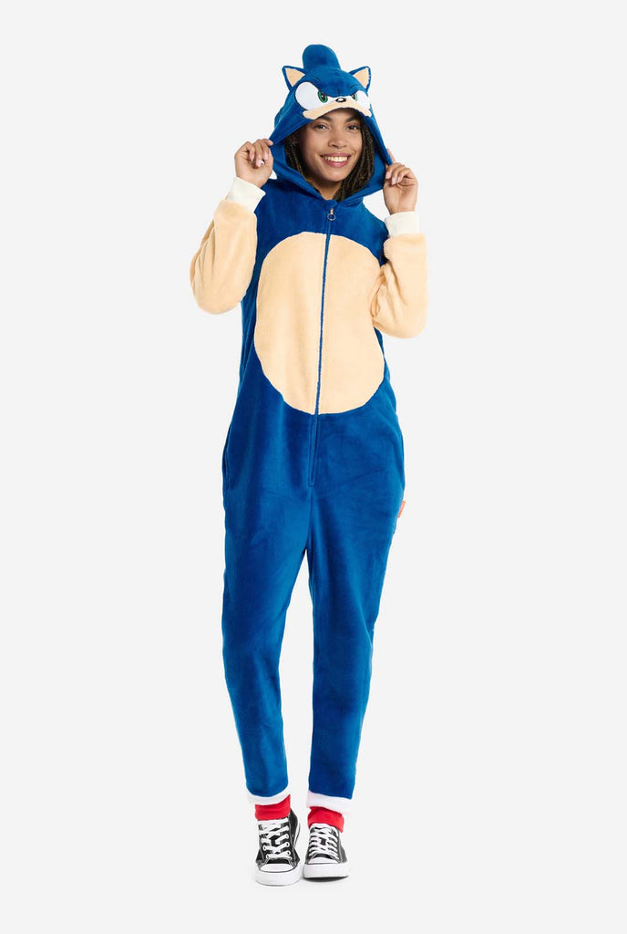 A woman is dressed in a cozy, blue Sonic the Hedgehog adult onesie, featuring soft beige patches on the front and sleeves to replicate Sonic's iconic look. The onesie has a hood designed with Sonic's recognizable face, including large green eyes, blue spikes, and pointy ears. The front zipper closure ensures a comfortable fit, and the woman is holding the hood. Her expression is calm and relaxed. This Sonic the Hedgehog onesie is ideal for cosplay, lounging, or themed events.