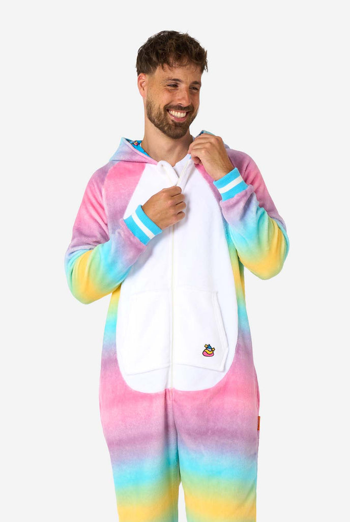 A man wearing a colorful adult onesie designed as a rainbow unicorn. The onesie features a vibrant gradient of pastel colors, a golden horn, and playful unicorn details, perfect for cozy lounging or costume parties. The hood is lying flat on his back.