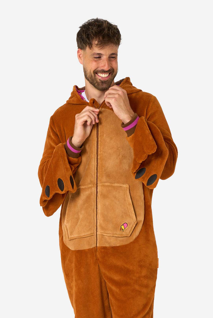 A cheerful man wearing a brown adult onesie designed as a walrus. The onesie features a hood with fangs, stitched gloves resembling paws, and playful walrus details, perfect for cozy lounging or costume parties. The hood is down on his back.