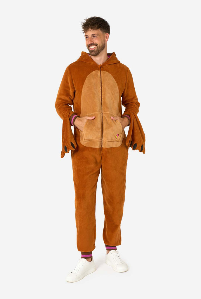 A cheerful man wearing a brown adult onesie designed as a walrus. The onesie features a hood with fangs, stitched gloves resembling paws, and playful walrus details, perfect for cozy lounging or costume parties. Full body image and the man holding his hand in his pockets.