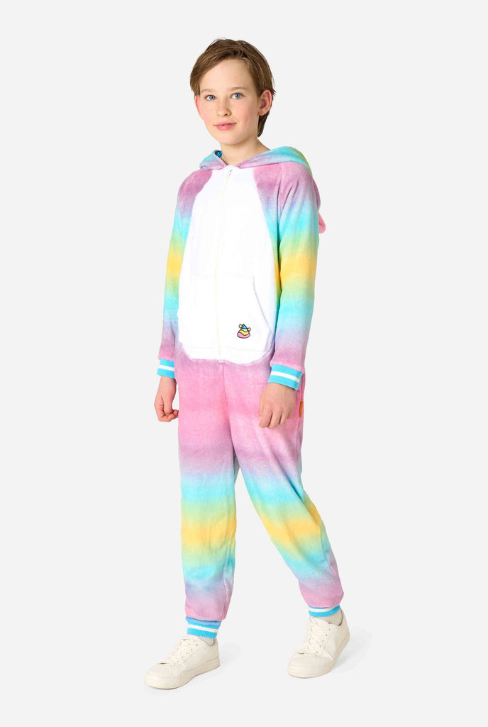 A smiling kid is wearing a white, pink, blue and yellow kids onesie designed as a unicorn. The Unicorn onesie features a hood resembling a unicorn head and other playfull details. Full body image with the hood down.