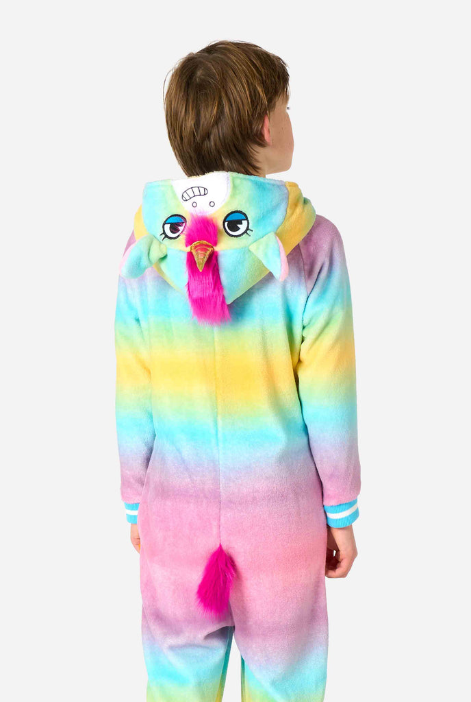 A smiling kid is wearing a white, pink, blue and yellow kids onesie designed as a unicorn. The Unicorn onesie features a hood resembling a unicorn head and other playfull details, View from the back with the hood down.