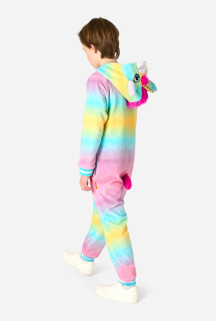 A smiling kid is wearing a white, pink, blue and yellow kids onesie designed as a unicorn. The Unicorn onesie features a hood resembling a unicorn head and other playfull details. Full body side view, with the hood down.