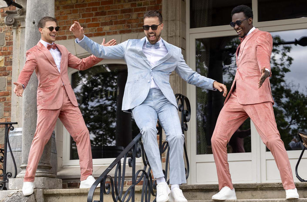 3 men on a line on a stair in front of a house wearing velvet tuxedos. The two outside men wearing a pink velvet tuxedo and the man in the middle wearing a blue velvet tuxedo.