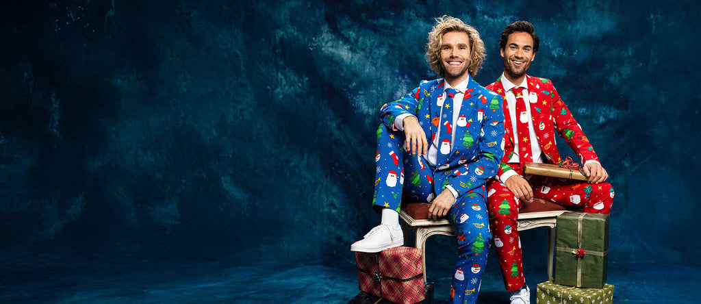 Two man sitting on a bench in front of a blue curtain wearing a Christmas suit. Left man wearing a blue Christmas suit and the right man wearing red Christmas suit.