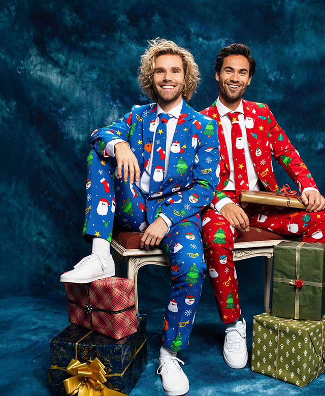 Two man sitting on a bench in front of a blue curtain wearing a Christmas suit. Left man wearing a blue Christmas suit and the right man wearing red Christmas suit.